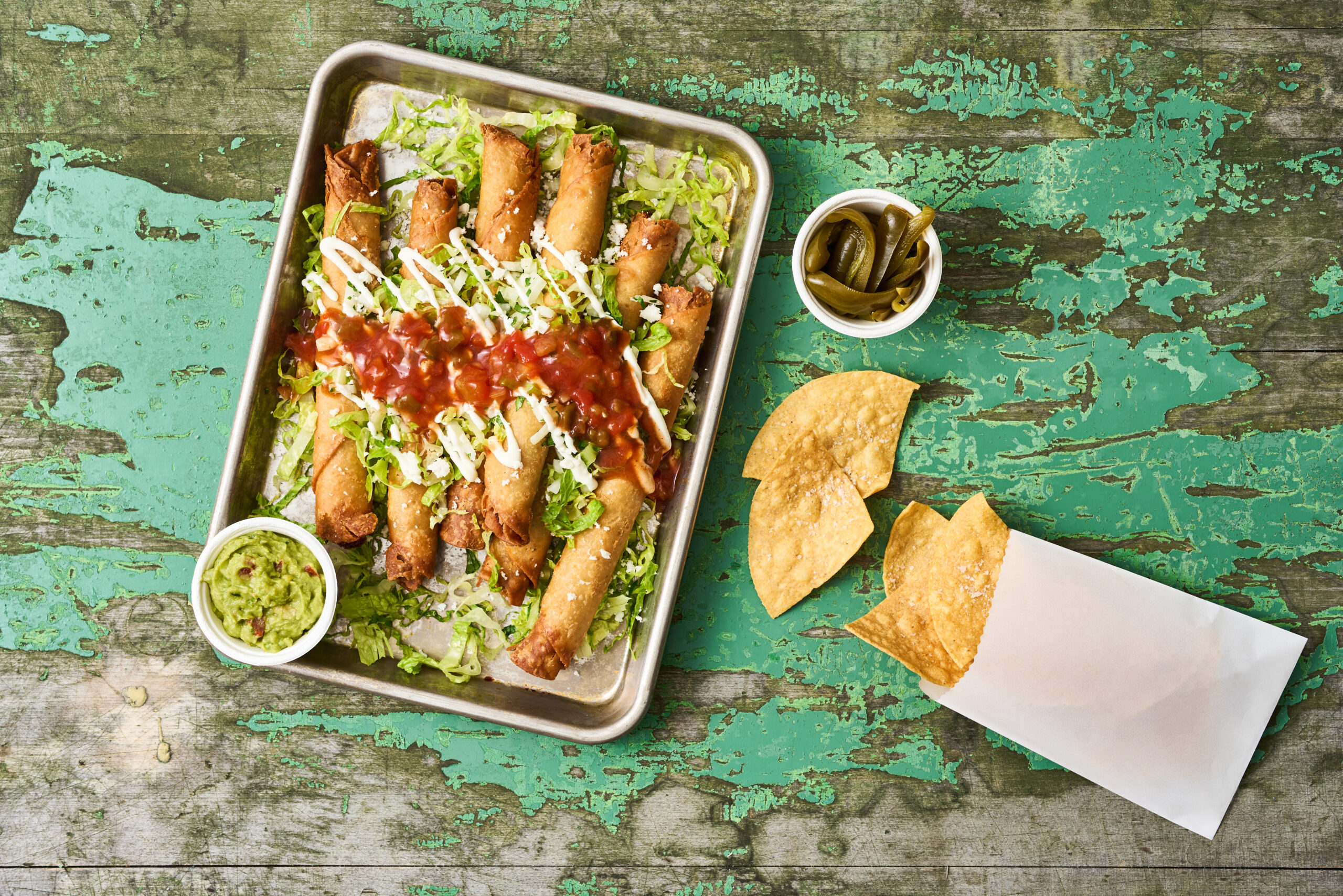 Top down shot of beef taquitos drizzled with Embasa Salsa Roja. Chips, guacamole, and veggies on the side.