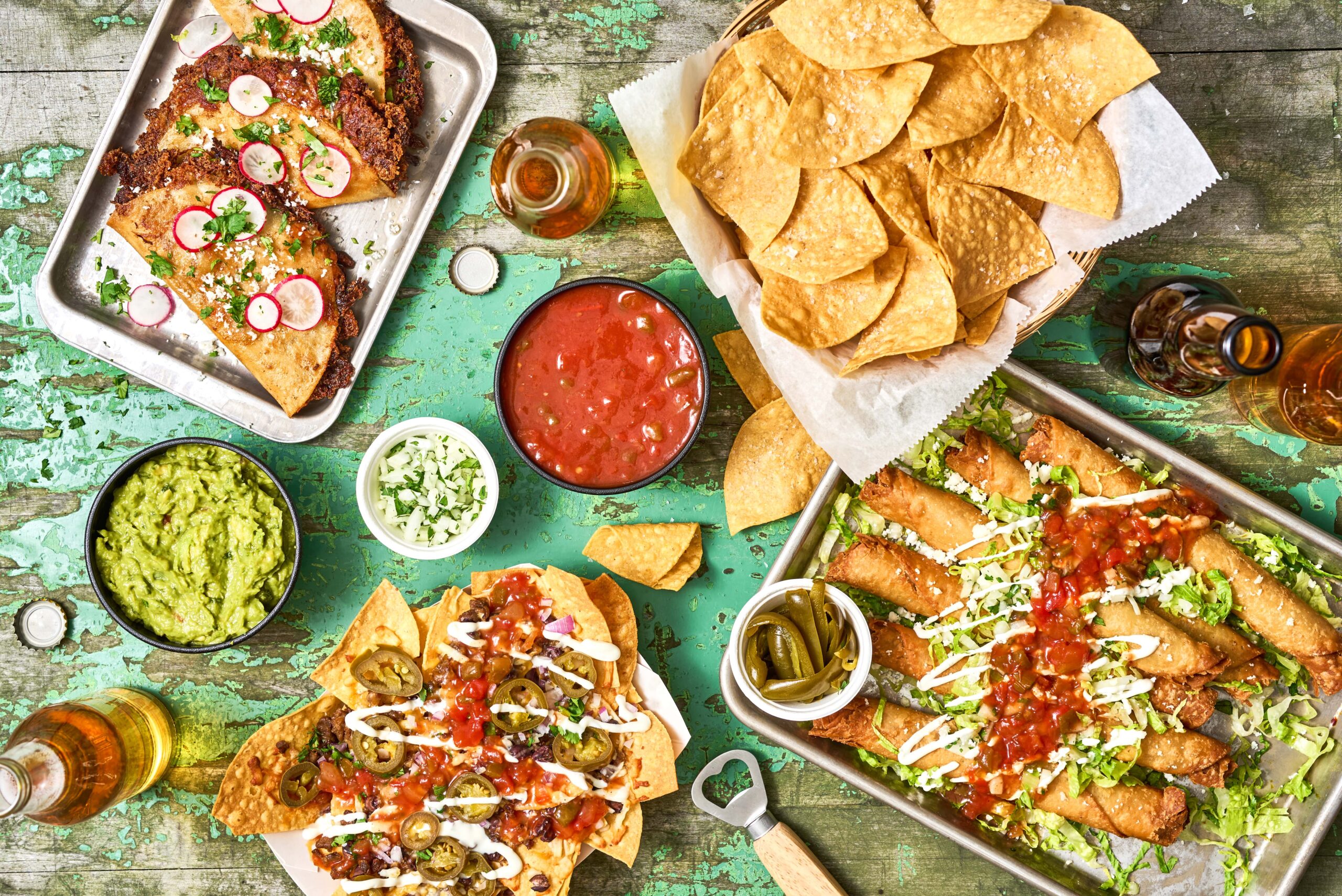 Top down shot of a family style table featuring tacos, nachos, taquitos, and chips with EMBASA Salsa Roja, Guacamole, diced onions, cilantro, and some drinks