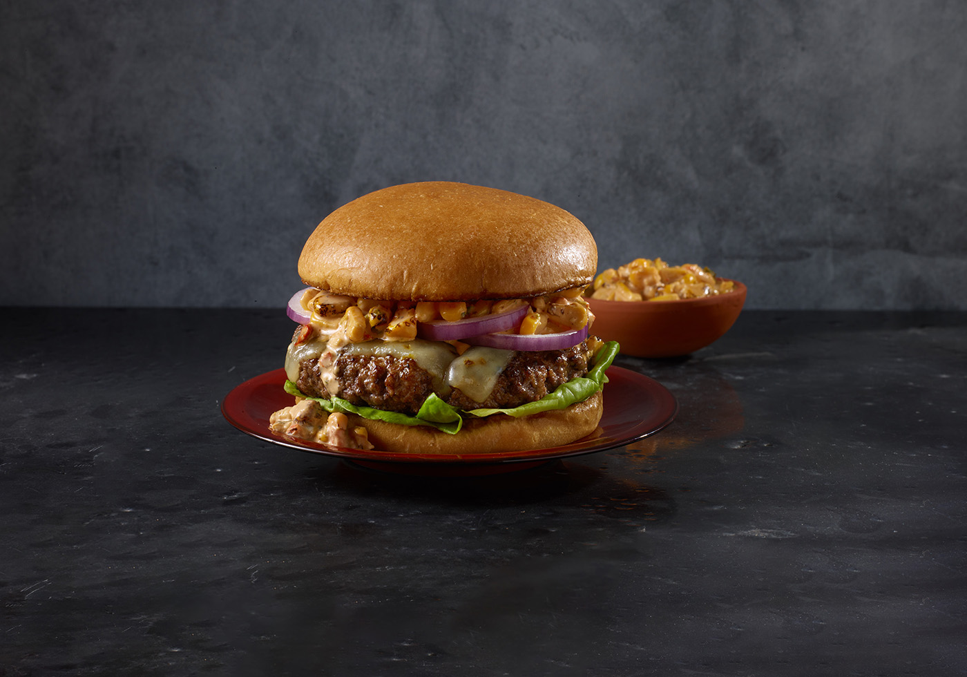 Burger with HERDEZ® Mexican Street Corn as a condiment. Orange bowl of street corn in the background.